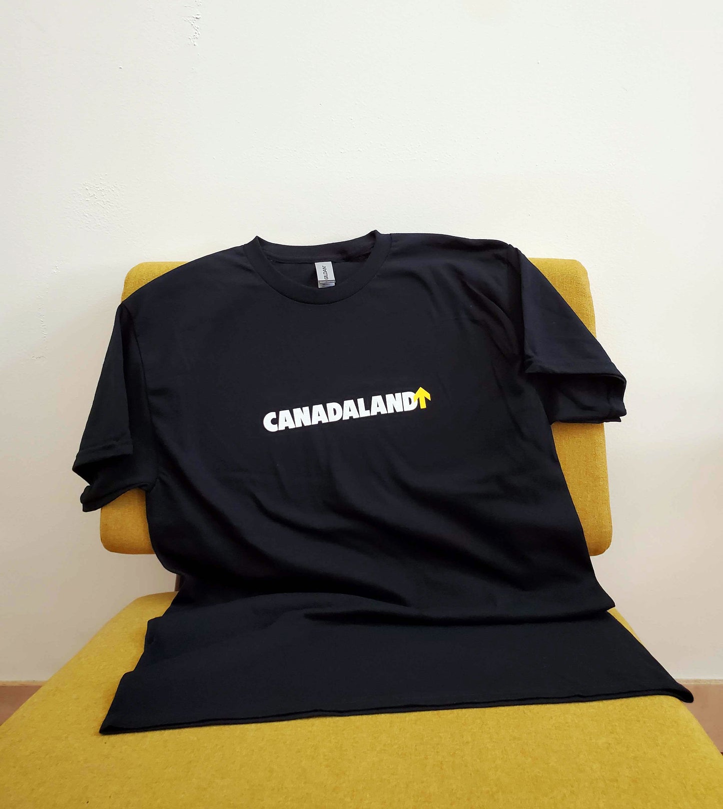 A black Gildan t-shirt, in 100% heavy weight cotton, with the Canadaland logo horizontal across the chest in white text and a yellow arrow.