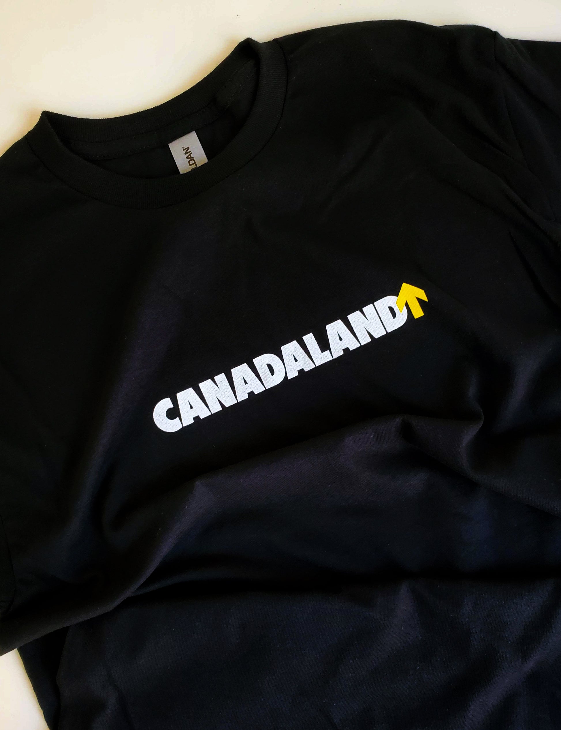 A black Gildan t-shirt, in 100% heavy weight cotton, with the Canadaland logo horizontal across the chest in white text and a yellow arrow.